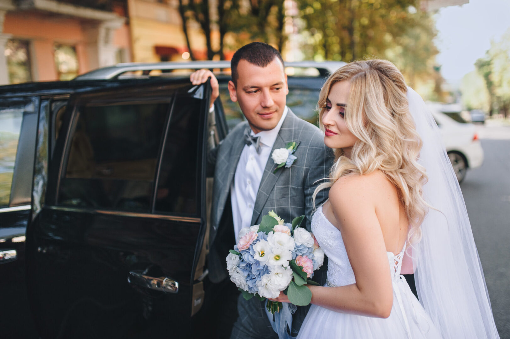Beautiful and stylish newlyweds stand in the city on the background of the car. A stylish bridegroom, leaning against the door of the car, looks at a sweet bride with curly hair.